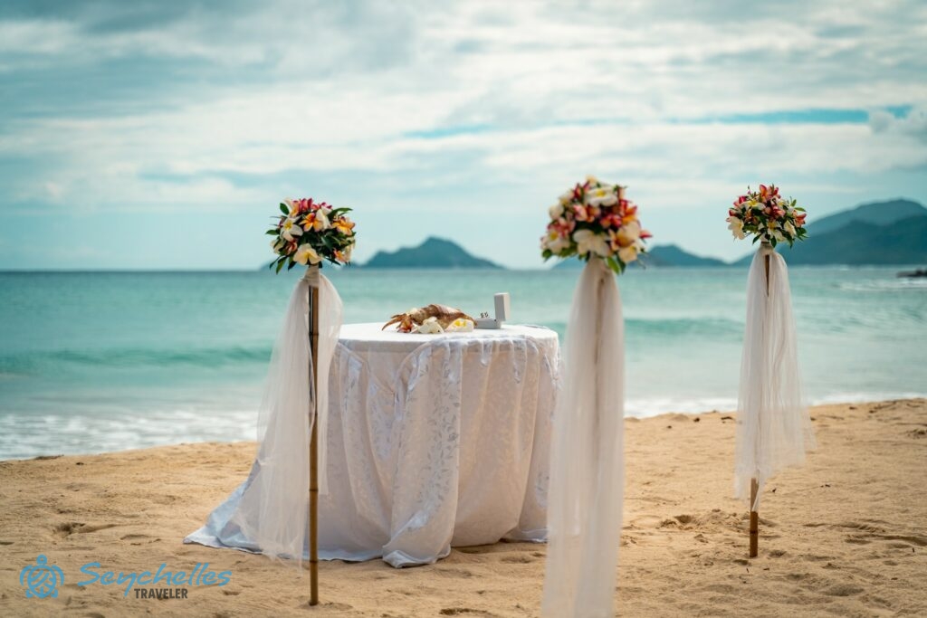 Színes virágokkal és fehér muszlinnal díszített bambusz oszlopokból álló bevonuló folyosó és asztal Anse Louis parton.
