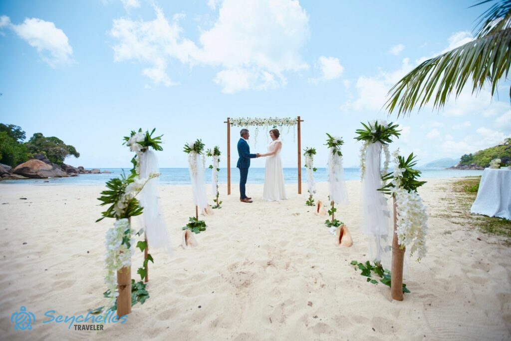 Fehér művirágokkal díszített bevonuló folyosó és a friss házasok a szögletes boldogságkapu előtt.