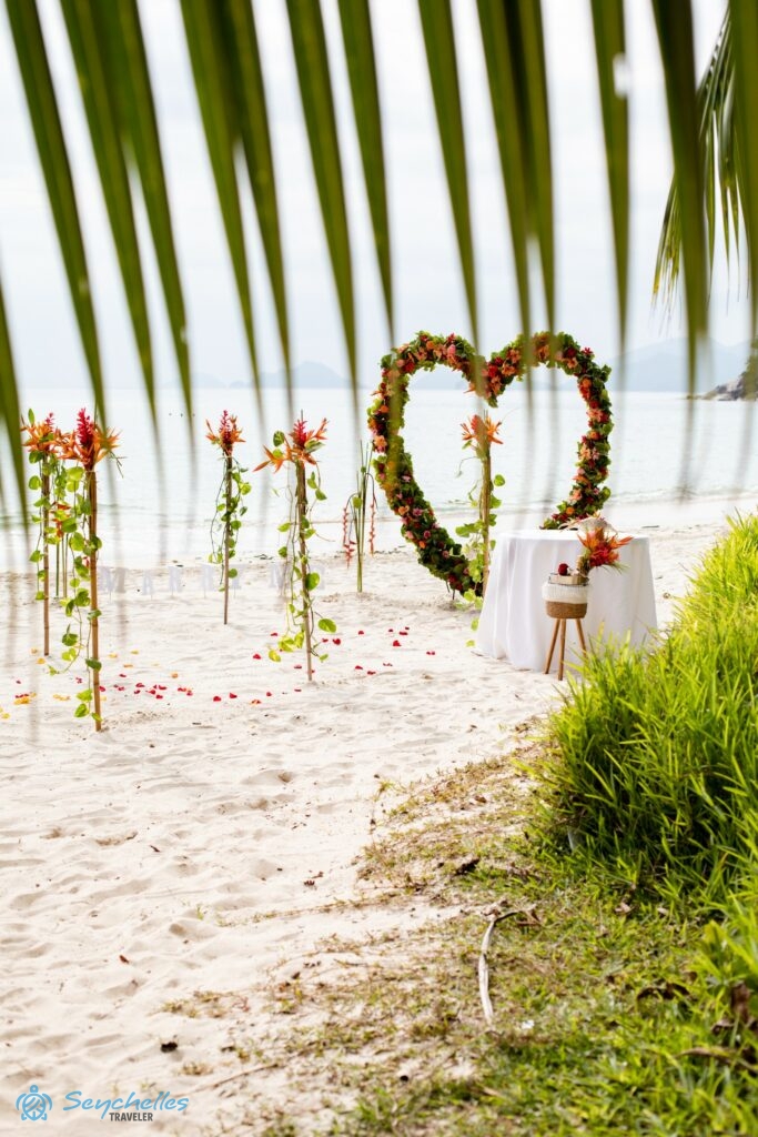 Szív alakú boldogságkapu és feldíszített bevonuló folyosó esküvői asztallal, pálmalevél mögül fényképezve Anse Louis parton.