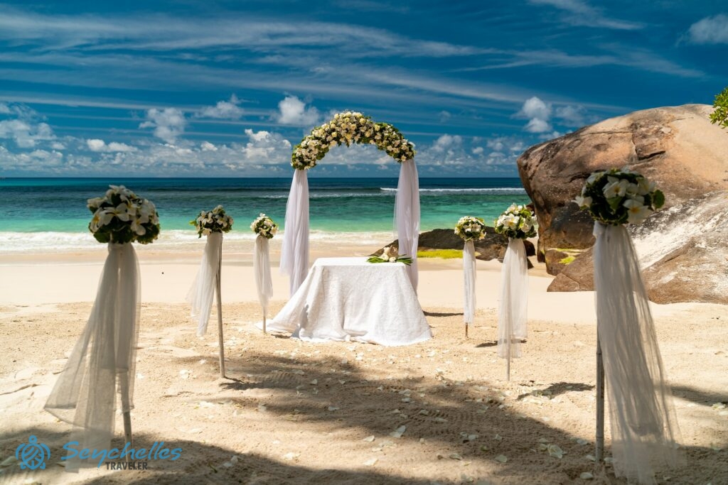 Fehér virágszirmokkal felszórt bevonuló folyosó, esküvői asztal dekorációval és íves boldogságkapu Baie Lazare tengerparton.