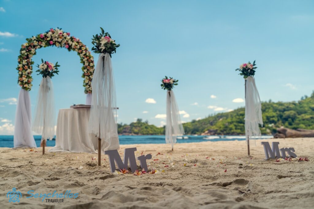 Virágokkal és muszlinnal díszített bevonuló folyosó, íves boldogságkapu és esküvői asztal Baie Lazare tengerparton.