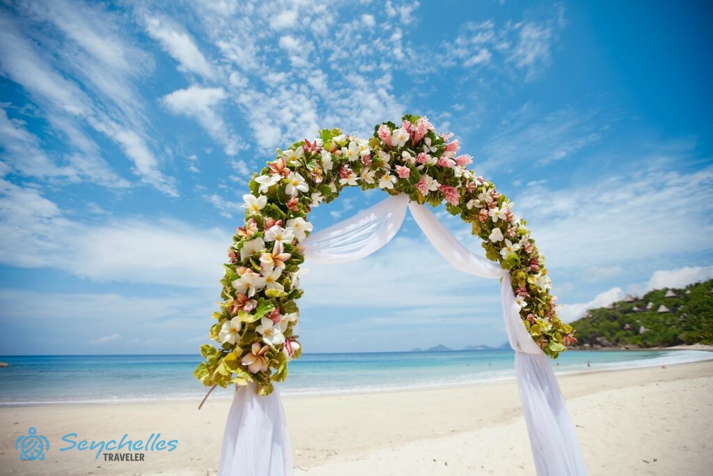 Frangipáni és gyömbér virágokkal sűrűn díszített, fehér muszlinnal bevont, íves alakú boldogságkapu Anse Louis parton.