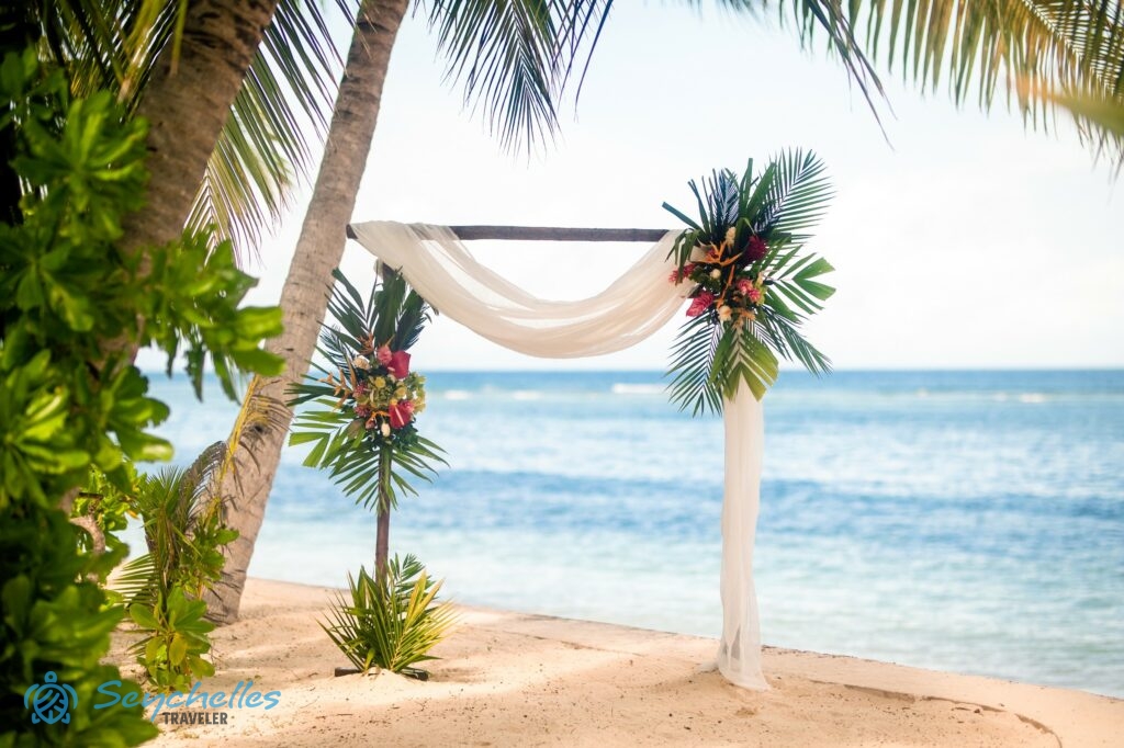 Színes virágokkal, fehér muszlinnal és pálmalevelekkel díszített szögletes boldogságkapu La Digue-en.