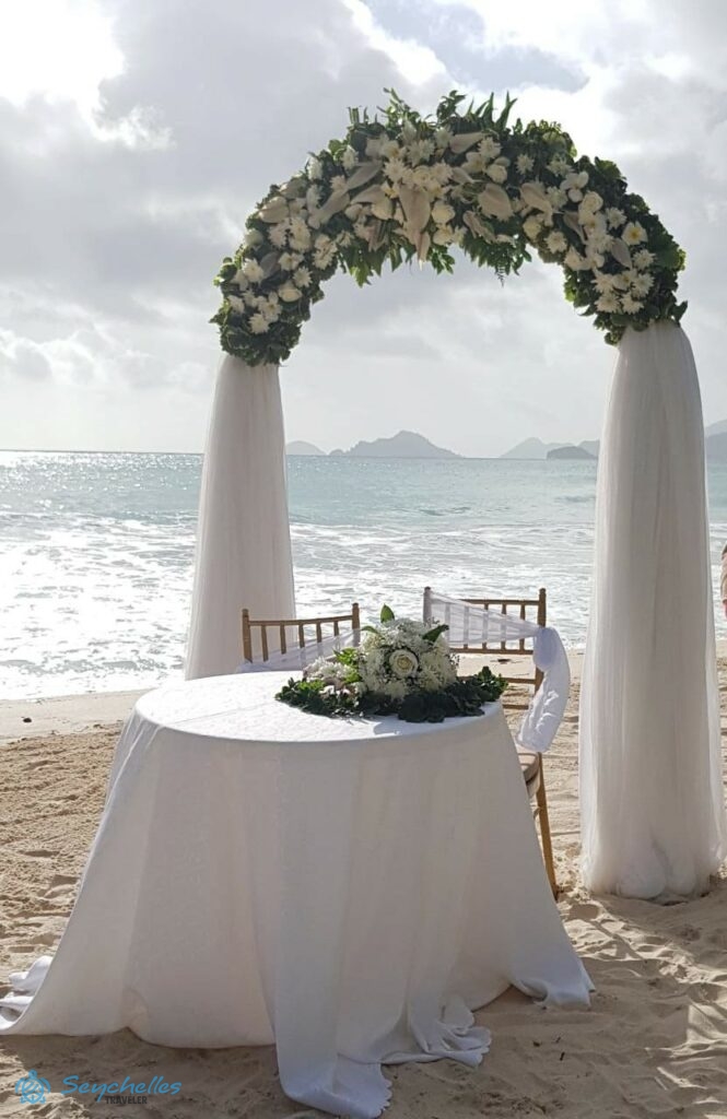 Esküvői asztal, íves boldogságkapu a közepén gazdagon díszítve fehér virágokkal, illetve az oszlopain sűrű, fehér fátyollal.