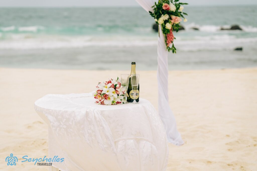 Esküvő asztal virágcsokorral, egy üveg habzóbor két pohárral, a háttérben boldogságkapuval North-East Point-on, Mahén.