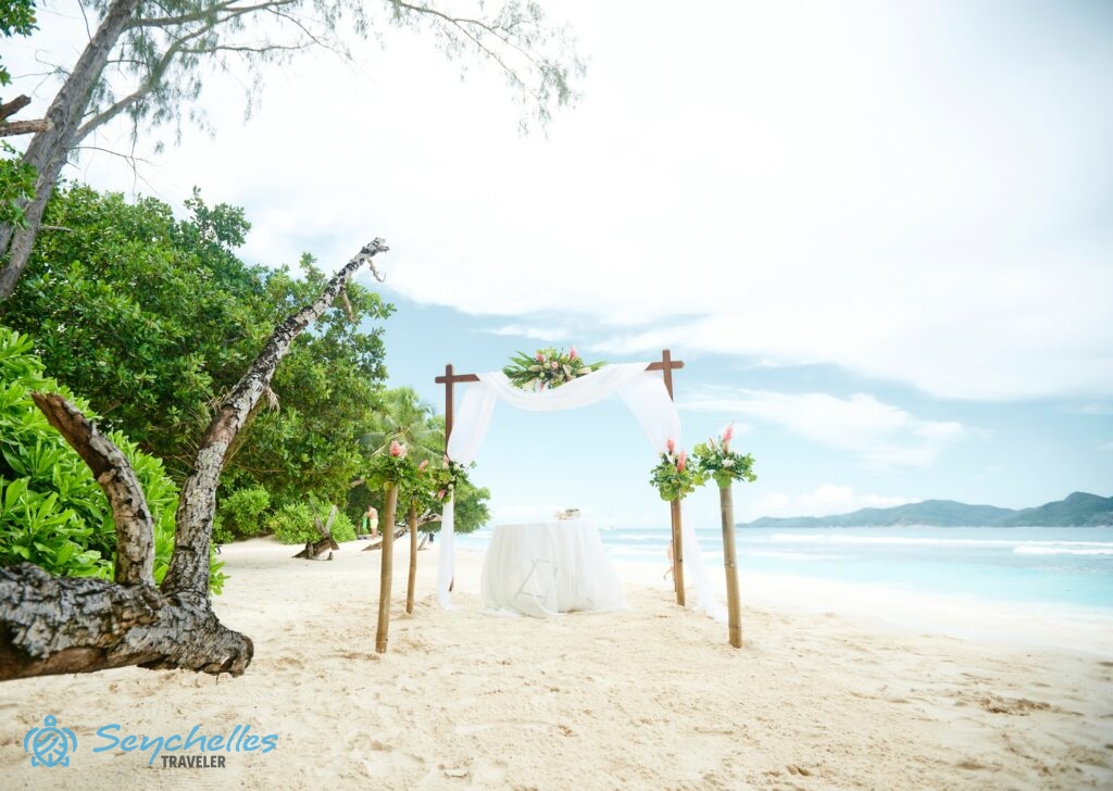 Szögletes boldogságkapu középen virág dekorációval, előtte négy oszloppal, melyeken virágcsokrok, esküvői asztal.