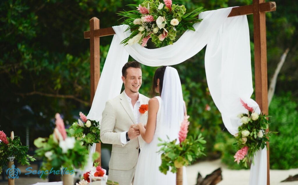 Fiatal pár a szögletes boldogságkapu előtt, körülöttük virág dekoráció Anse Severe tengerparton.