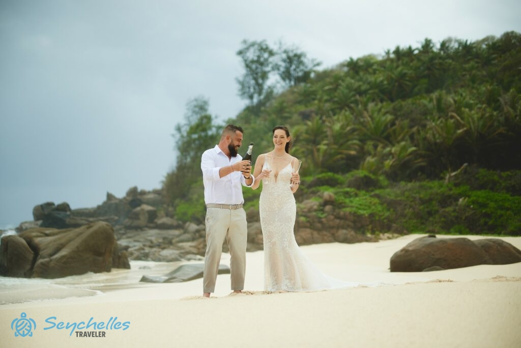 Mahén, Anse Bazarca esküvő helyszínen egy fiatal pár áll a tengerparton pezsgővel és két pohárral a kezükben.