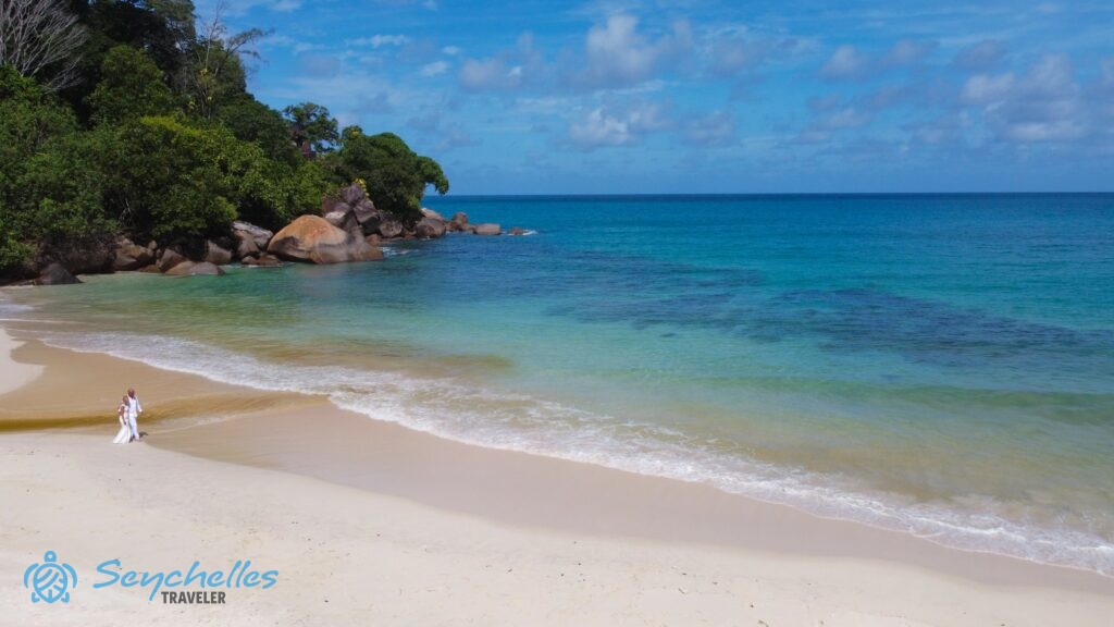 Drónfelvétel fehérbe öltözött friss házasokról, amint Anse Louis esküvői helyszínen a homokban sétálnak, előttük az óceán.