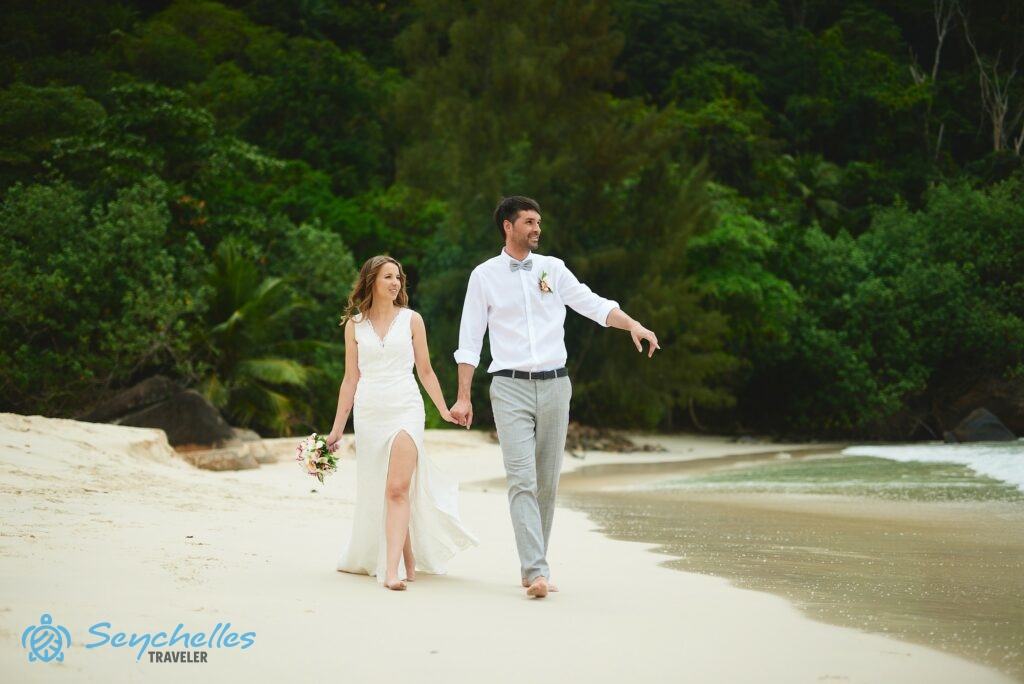 Friss házasok figyelik az óceánt Mahén, Anse Louis tengerpartján sétálva, mögöttük sűrű trópusi vegetáció.