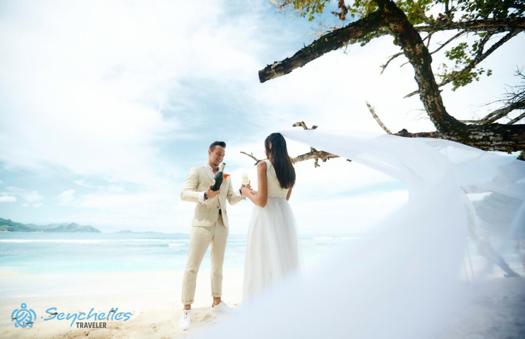 A menyasszony hosszú fátylát meglobbantja a szél miközben a vőlegény pezsgőt tölt neki Anse Severe tengerparton, La Digue-en.