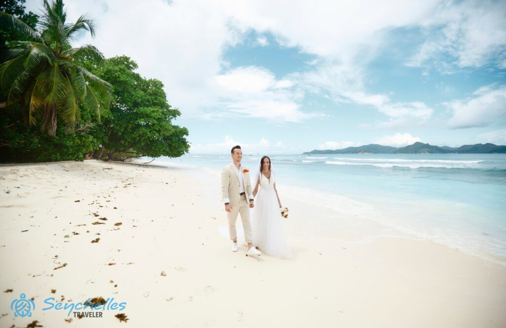 A fiatal pár Anse Severe tengerparton sétál, a homok fölé trópusi fák nyúlnak be, bal oldalukon pedig az óceán.