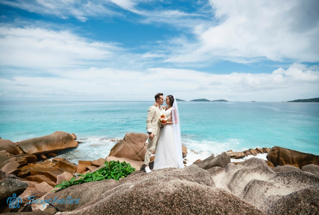 Házaspár áll Anse Severe gránitszikláin La Digue-en, a háttérben a kristálytiszta óceán és a fehér felhőkkel tűzdelt égbolt.