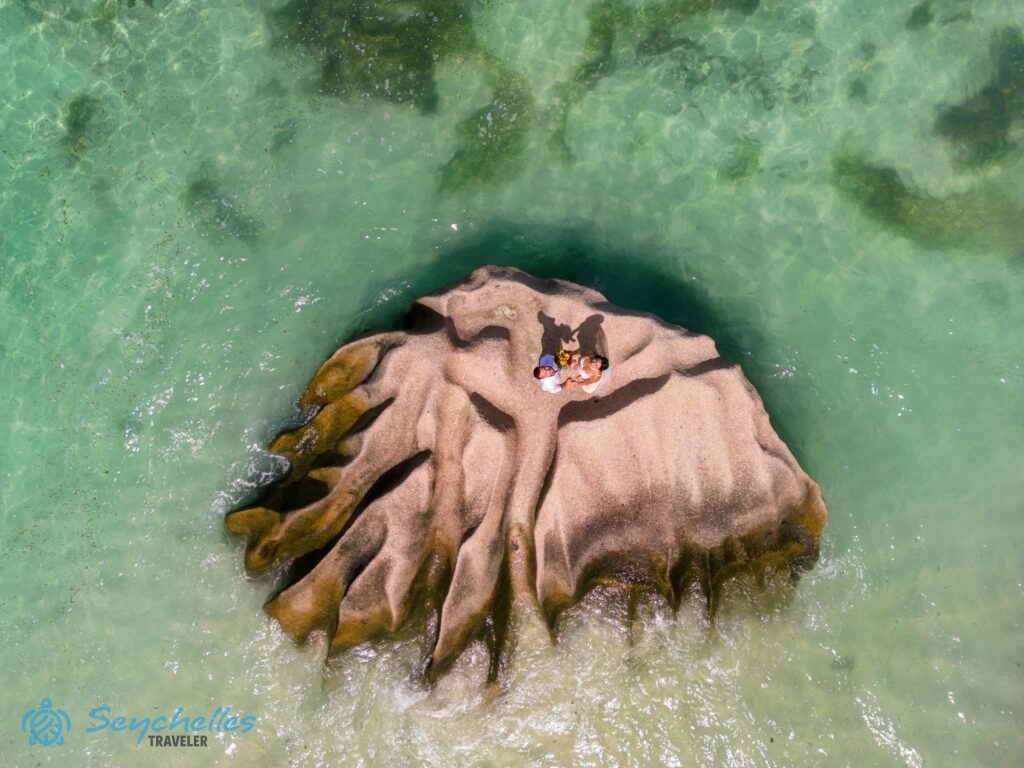 Légi felvétel a kristálytiszta óceánból kimagasodó sziklán álló házaspárról Anse Source D'Argent parton La Digue-en.