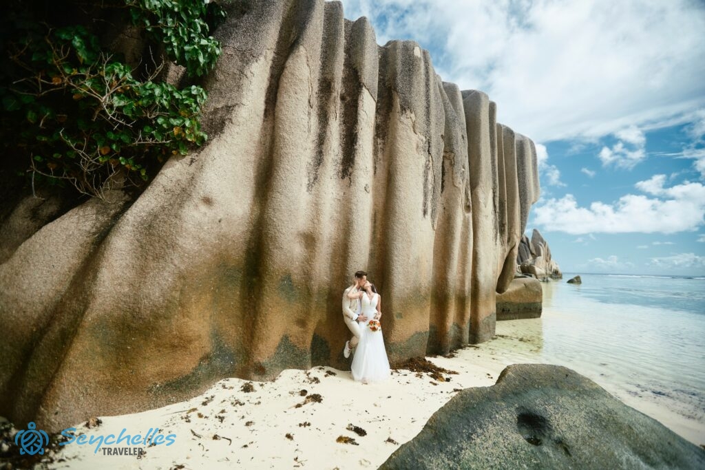 Házaspár Anse Source D'Argent parton, az átlátszó óceán közelében egy hatalmas, bordázott sziklafalnak támaszkodva.