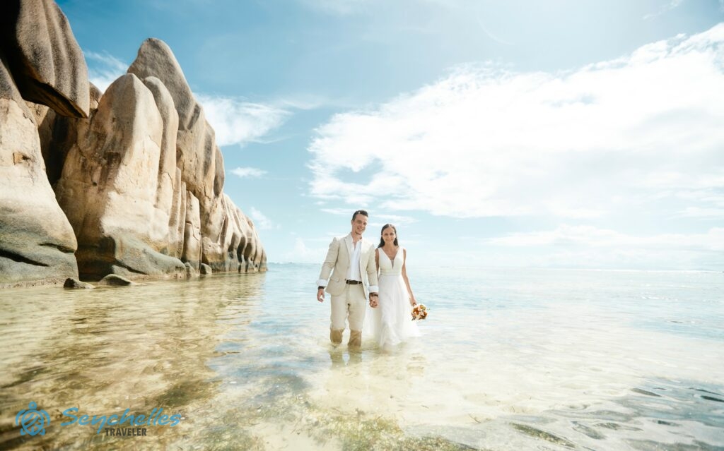 Az ifjú pár egy vízből kiemelkedő szikla szélén áll Anse Source d'Argent tengerparton, a távolban Praslin látszódik.
