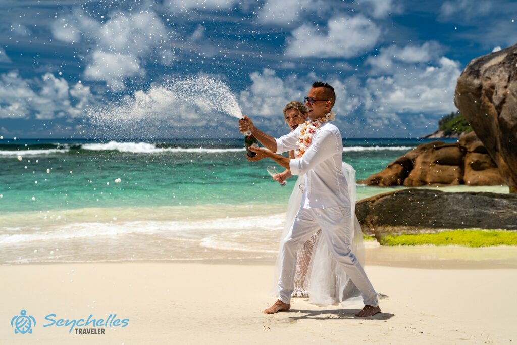 Baie Lazare tengerparton a vőlegény kezében felbontás után fröcsköl a pezsgő, a menyasszony nevetve figyeli.
