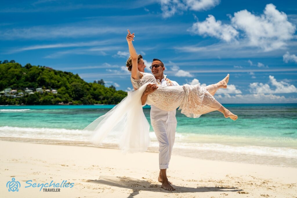 A vőlegény mosolyogva emeli karjaiban a menyasszonyt Baie Lazare tengerparton, mögöttük az óceán változó színű kékje.