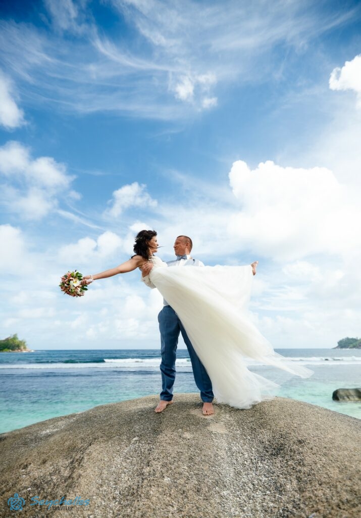 Baie Lazare tengerparton a Roche Copra szikla tetején a vőlegény a karjába veszi a menyasszonyt, mögöttük az óceán.