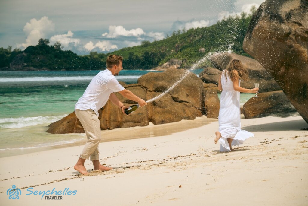 A vőlegény pezsgővel locsolja az előle menekülő menyasszonyt Baie Lazare tengerparton egy gránitszikla tövében.