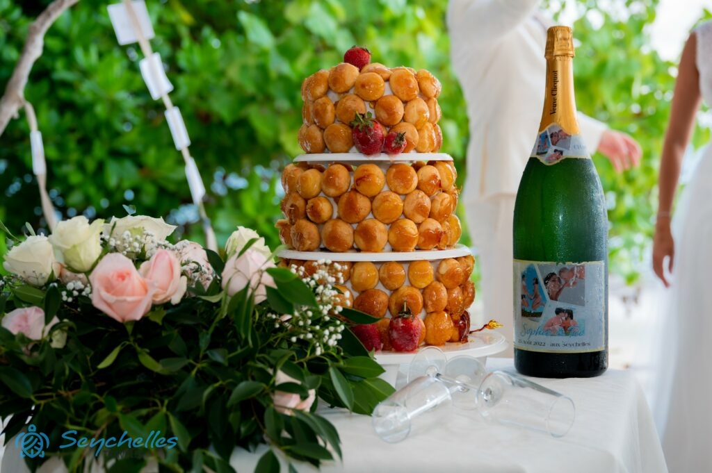 Három emeletes esküvői torta egy üveg habzóborral a párról készült képekkel, két pohár elfektetve és asztaldísz La Digue-en.