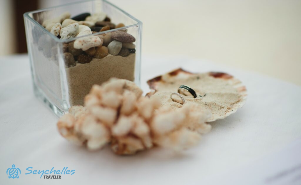 Homokkal töltött kagyló gyűrűtartó az asztalon La Digue-en, mellette díszítés korallból, homokból és kavicsokból.