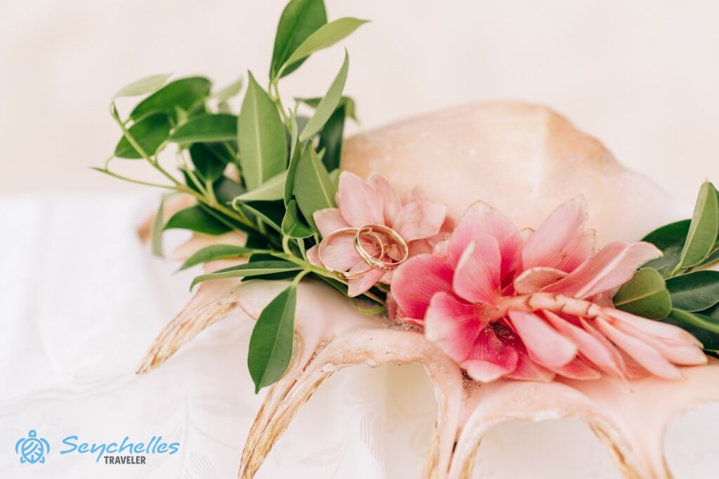 Rózsaszín gyömbér virágokkal és zöld ágacskákkal díszített csigaház gyűrűtartón pihenő gyűrűk Mahén.