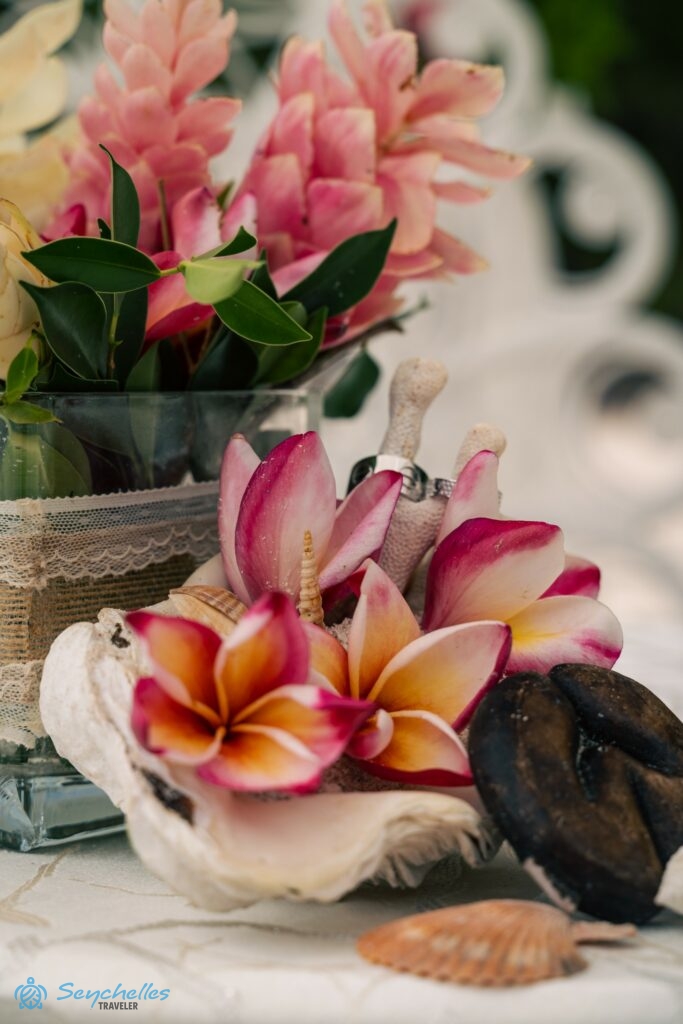 Óriáskagyló gyűrűtartó csigaházakkal, frangipáni virágokkal, korallokkal, és kis, fából faragott tengerikókusz dísszel.