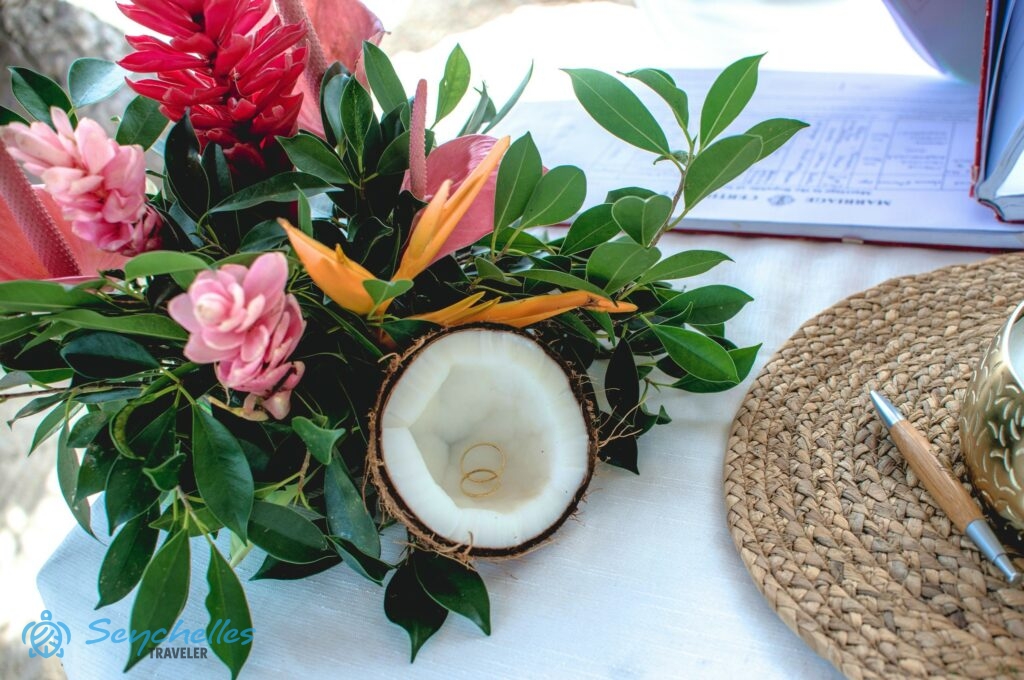 Asztali dísz gyömbér és papagájvirággal, előtte gyűrűtartó félbevágott, kókuszból, és benne pihenő gyűrűk Praslin-i esküvőn.