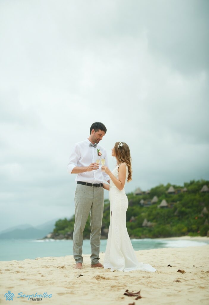 A friss házasok Anse Louis tengerparton a homokban állva habzóborral koccintanak.