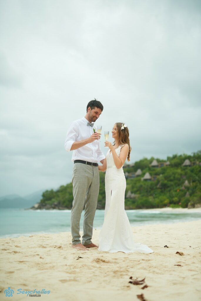 Az ifjú pár egymást átölelve, a kezükben pezsgőspohárral mosolyogva néz egymásra Anse Louis tengerparti esküvőn.