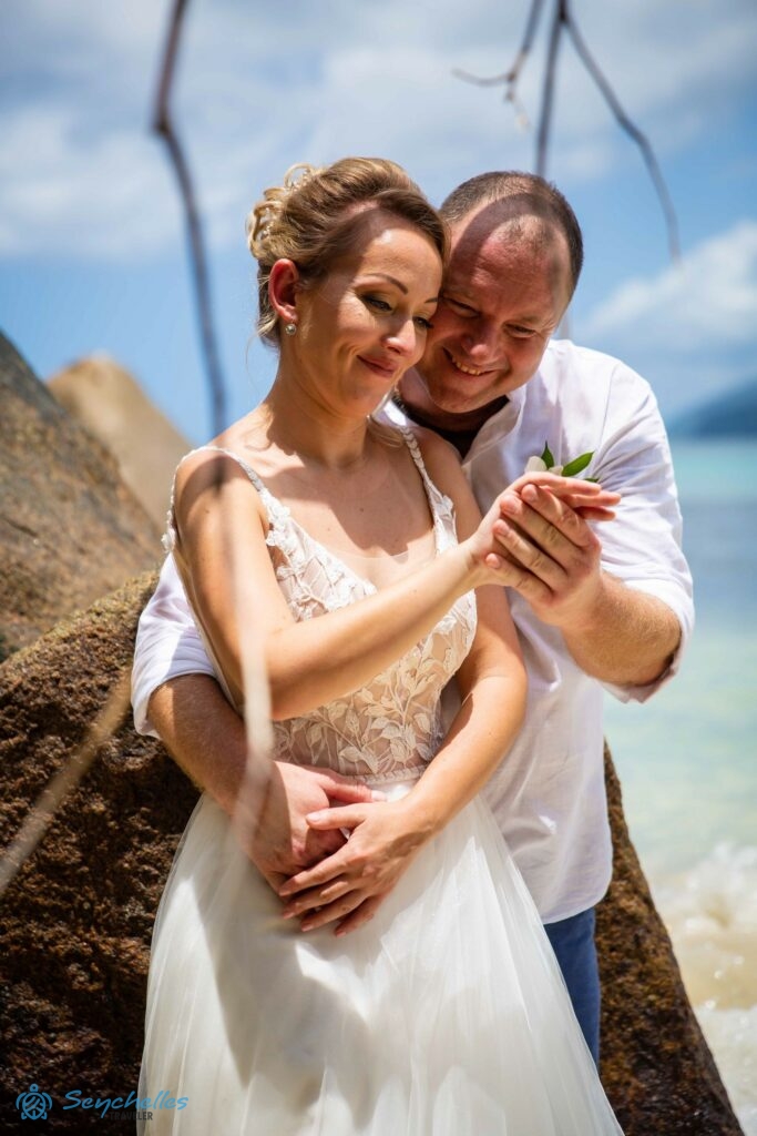 Feleség és férj mosolyogva nézik a karikagyűrűt a feleség ujján, amint egy szikla előtt állnak Anse Louis parton.