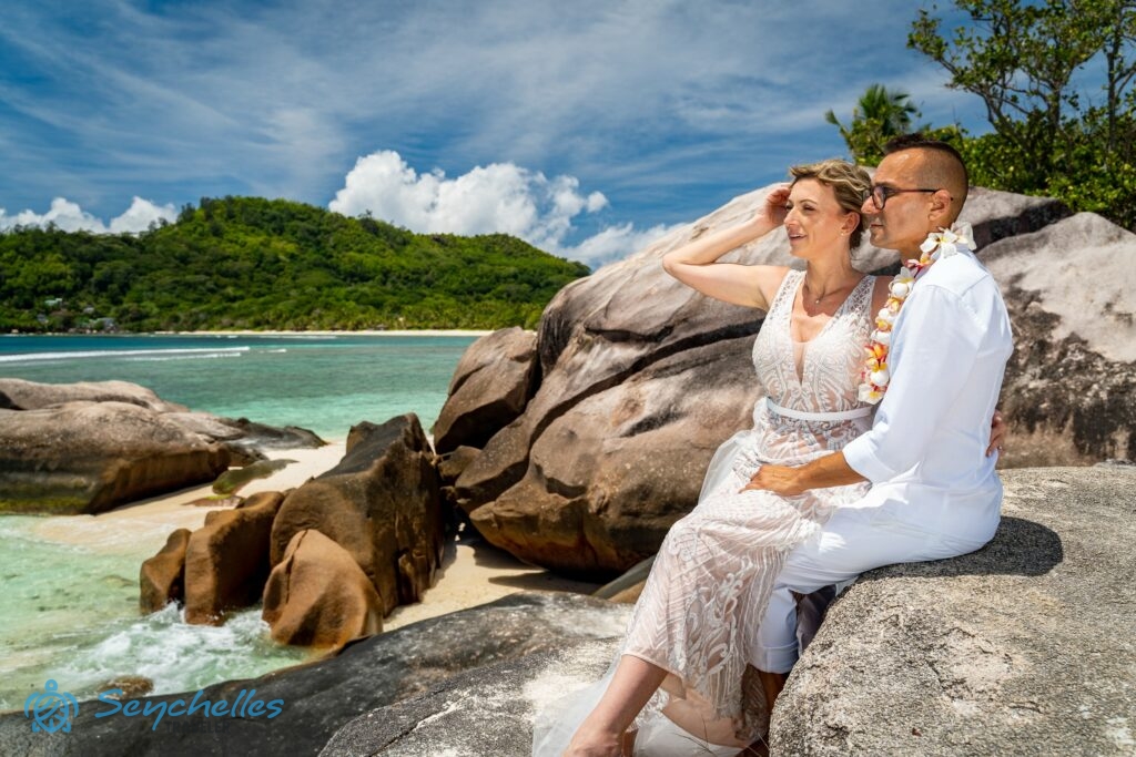 Gránitsziklák tetején ülő, fehérbe öltözött friss házasok a tengert nézik Baie Lazare tengerparton.