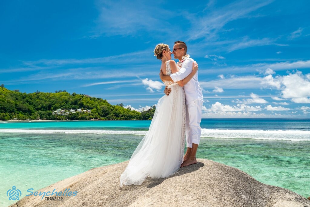Az újdonsült házaspár egy nagy gránitszikla tetején egymással szemben áll Baie Lazare tengerparton, mögöttük a ragyogó óceán.