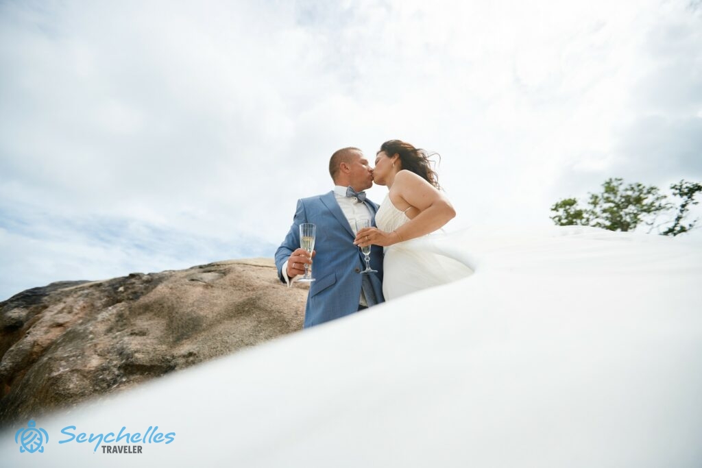 Friss házasok egy szikla előtt pezsgőspohárral a kezükben Baie Lazare tengerparton.