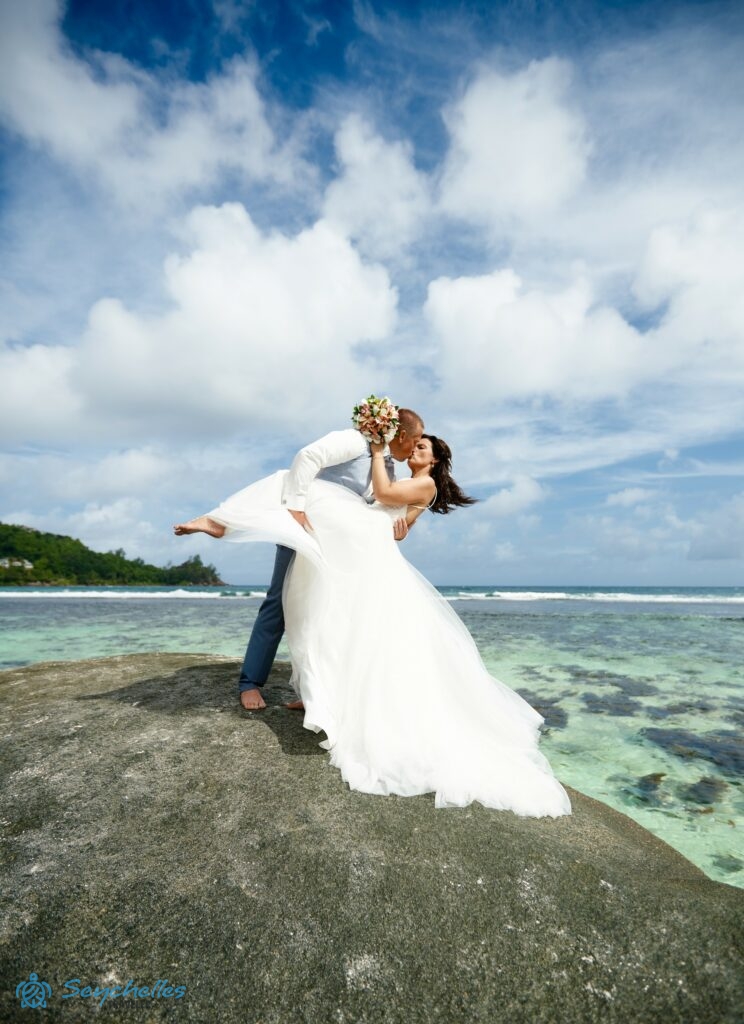 A férj felesége derekát és lábát tartva dönti hátra egy szikla tetején állva Baie Lazare tengerparton.