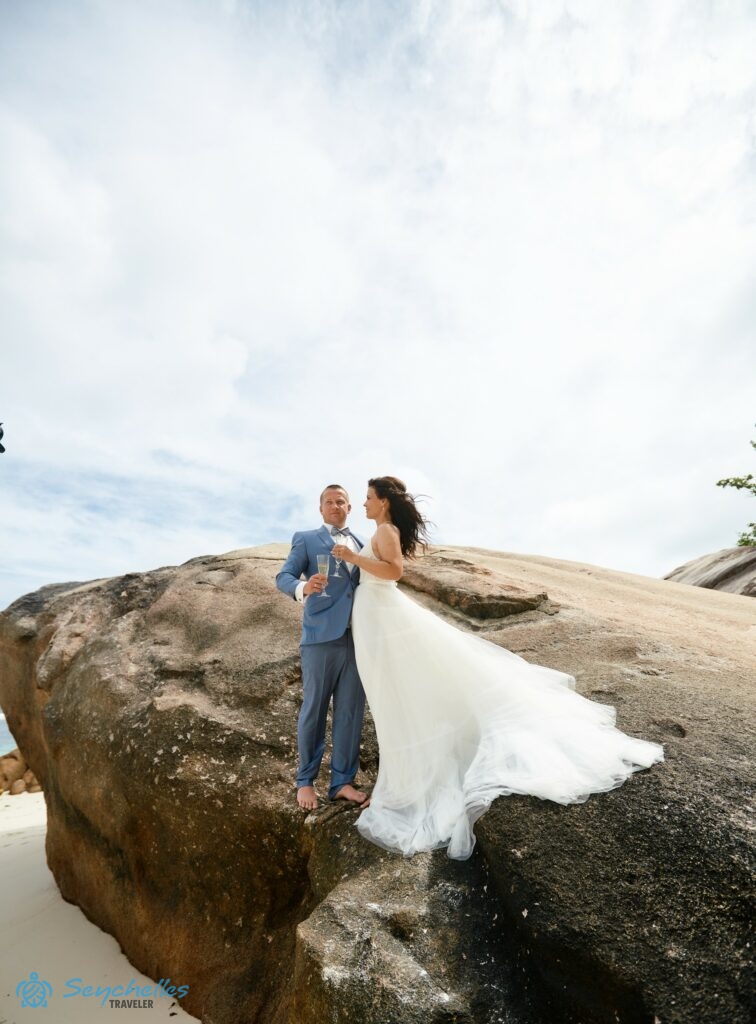 A friss házasok pezsgőspohárral a kezükben állnak egy szikla alján Baie Lazare tengerparton.