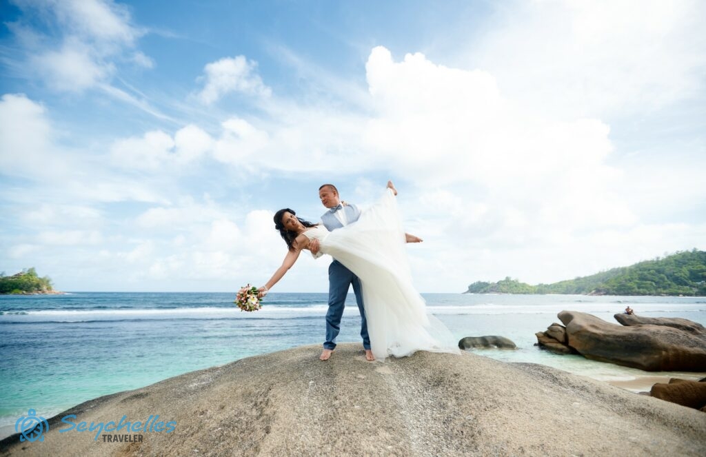 Esküvői fotó a menyasszonyt a karjában tartó vőlegényről egy szikla tetején Baie Lazare tengerparton.