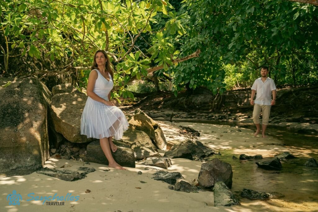 Esküvői fotó a sziklának támaszkodó feleségről és a felé sétáló férjről Baie Lazare partnál.