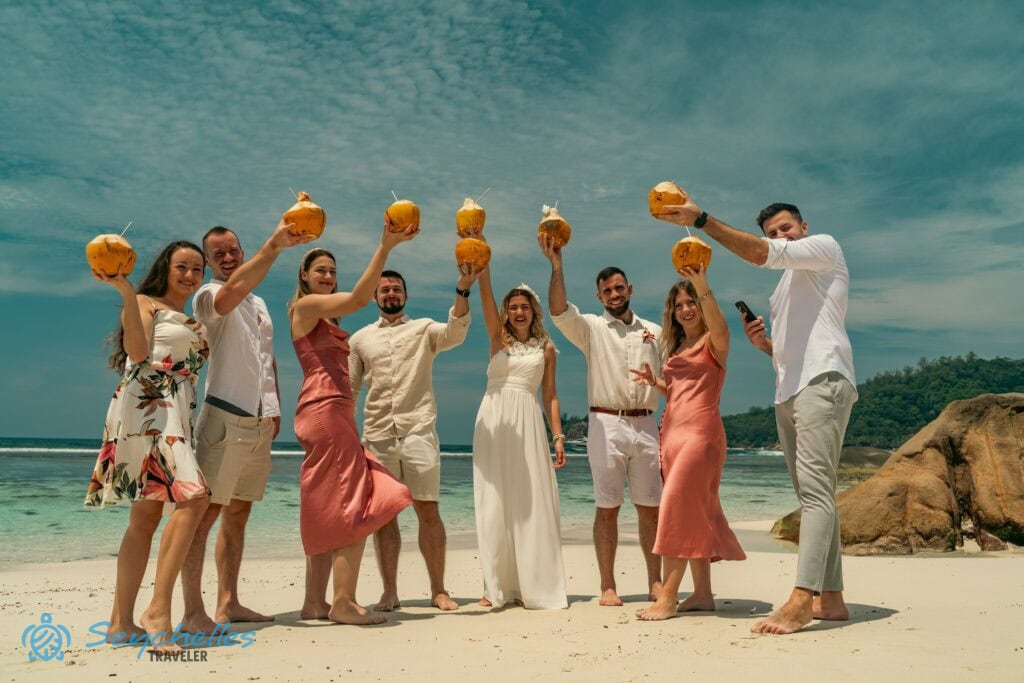 Esküvői fotó a friss kókuszitalokat magasba emelő házaspárról és barátaikról Baie Lazare tengerparton.