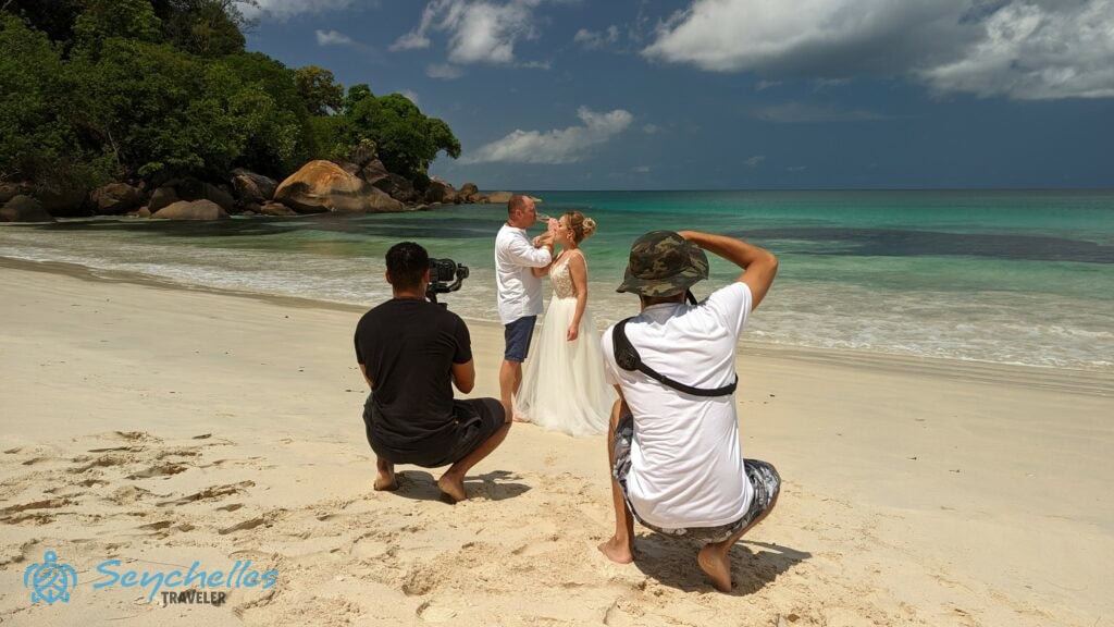 A pezsgőt fogyasztó friss házasok közelében fényképészek készítenek róluk esküvői felvételeket Anse Louis tengerparton.