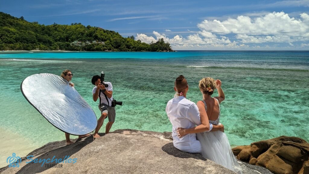 Az esküvői fotózás alatt a fényképész mellett álló Réka tartja a derítőlapot a sziklán ülő házaspár felé Baie Lazare parton.