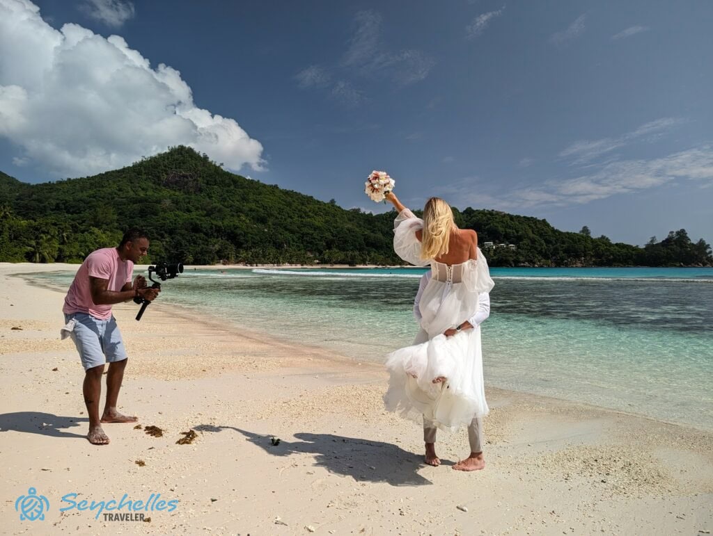 A vőlegény a magasba emeli a menyasszonyt, miközben a fotós képeket készít róluk Baie Lazare tengerparton.