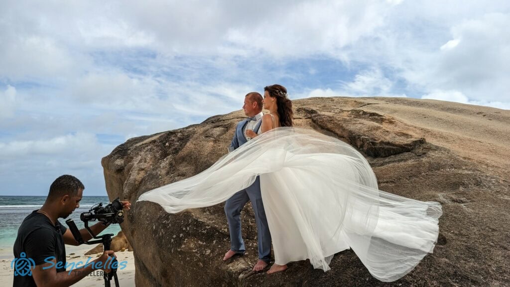 A fotós a menyasszony libbenő szoknyája mellől készít képeket a házaspárról Baie Lazare tengerparton egy sziklánál.