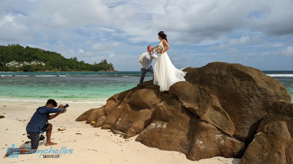 A fotós a homokban térdelve készít képeket a sziklán álló házaspárról Baie Lazare tengerparton.
