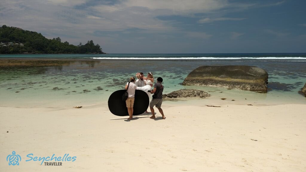 Az esküvői fotózáson Réka a derítőlapot tartja a tenger szélén állva, miközben képek készülnek a párról Baie Lazare parton.