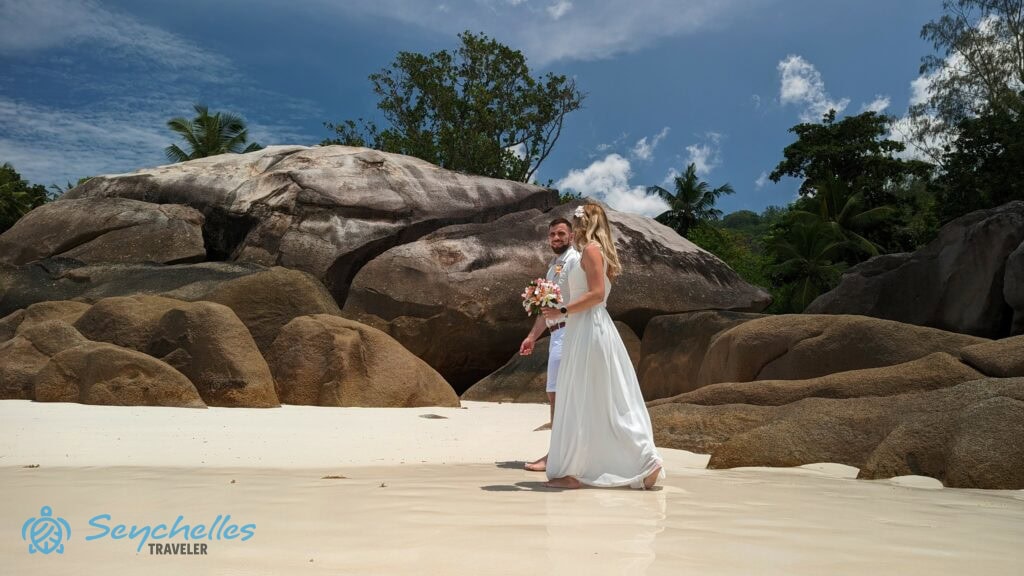 Esküvői werkfotó a homokban sétáló házaspárról Baie Lazare szikláival a háttérben.
