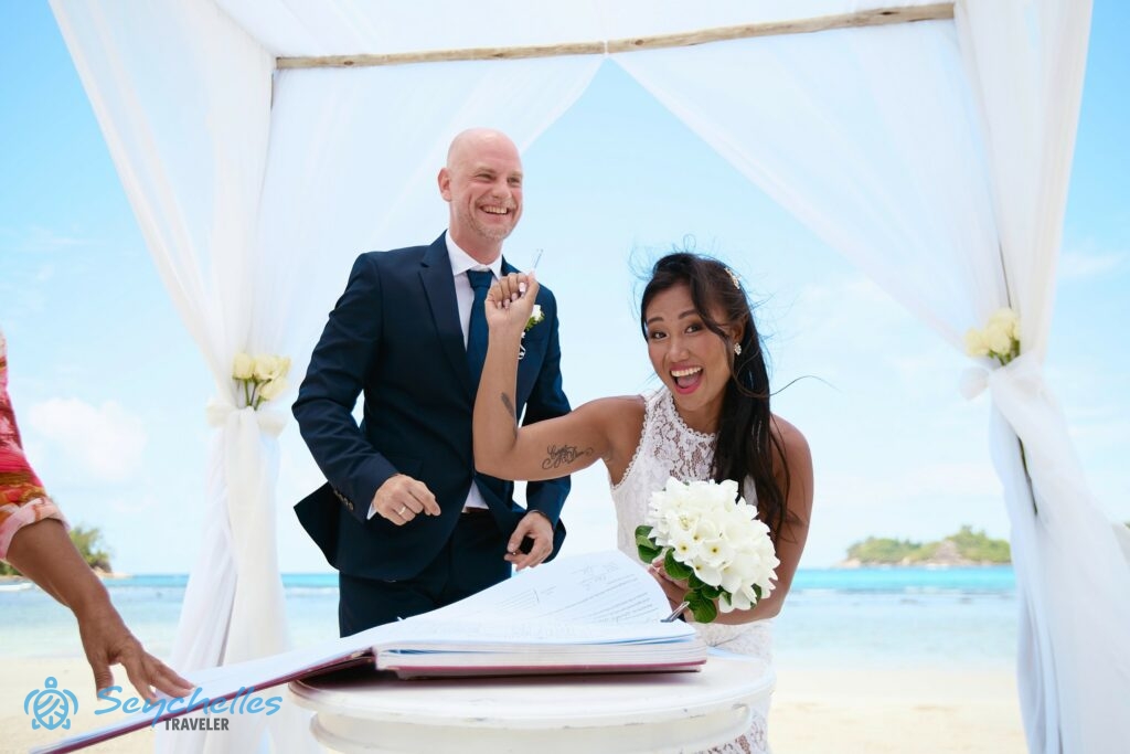 A pár a házassági anyakönyvi kivonat hivatalos példányát írja alá Baie Lazare parton az angol nyelvű hivatalos esküvőn.