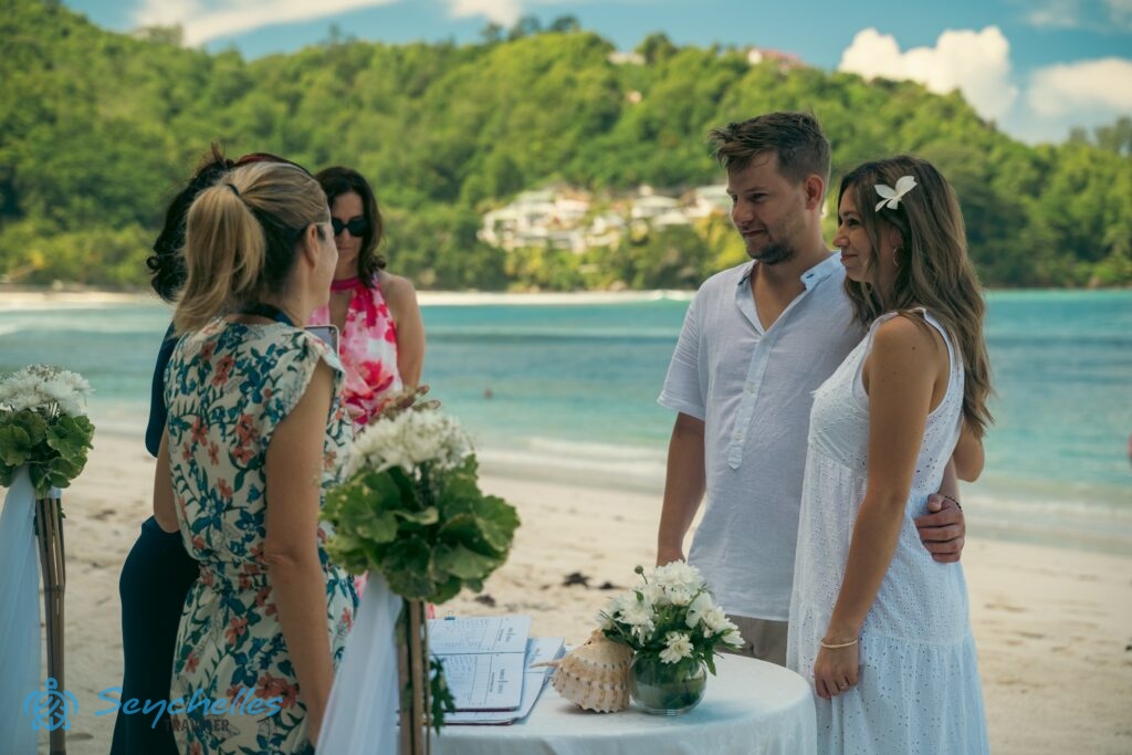 A pár a feldíszített asztalnál hallgatja Rékát és az anyakönyvvezetőt, Baie Lazare parton a magyar nyelvű, hivatalos esküvőn.