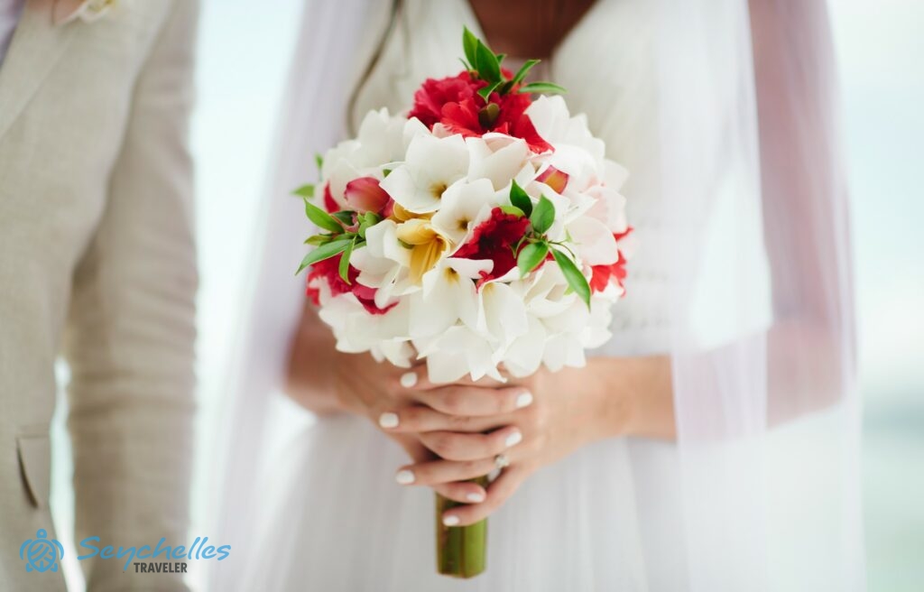 A menyasszony a fehér és piros virágokból készült, zöld levelekkel tűzdelt menyasszonyi csokrot tartja a kezében.